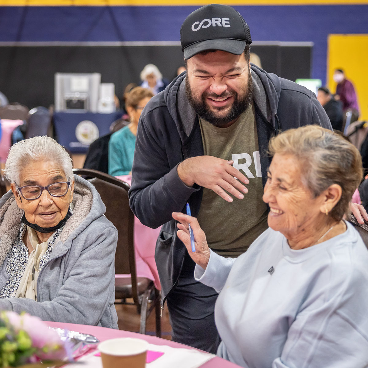 A male CORE staffer laughs with two elderly women at a table during a community health event.