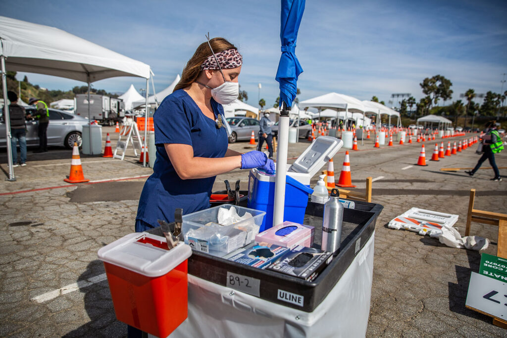 Nurse prepares vaccines at a mobile station in the middle of the Dodger Stadium parking lot. Needle disposal boxes, ice chests to keep vaccines cold, and other supplies lay on the stand.