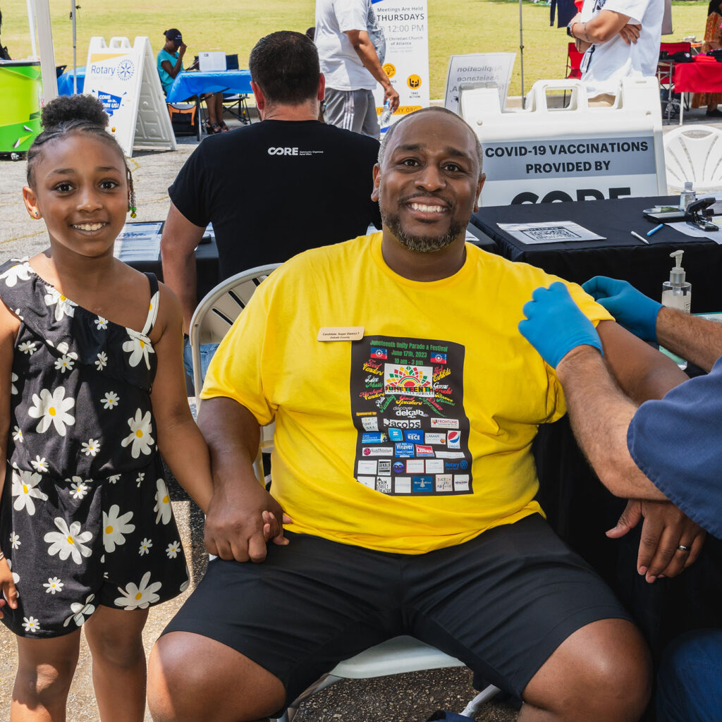 A father and his young daughter smile at the camera. The father is sitting in a chair after getting vaccinated at CORE's stand at a local community event in Georgia.