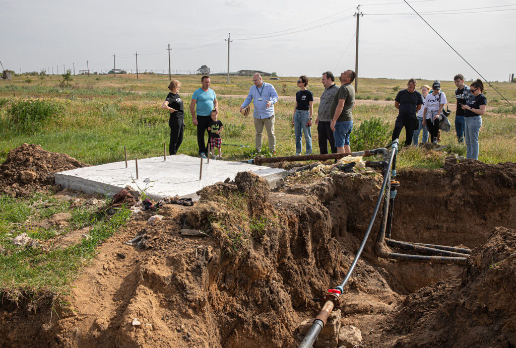 CORE staff, community leaders, and partners stand around ground construction to install pipe.