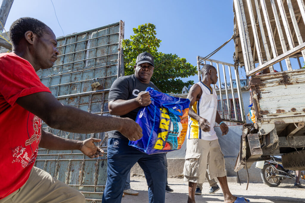 Clausel, Haitian man and CORE staff, lifts sack of food and hands it to another haitian man.