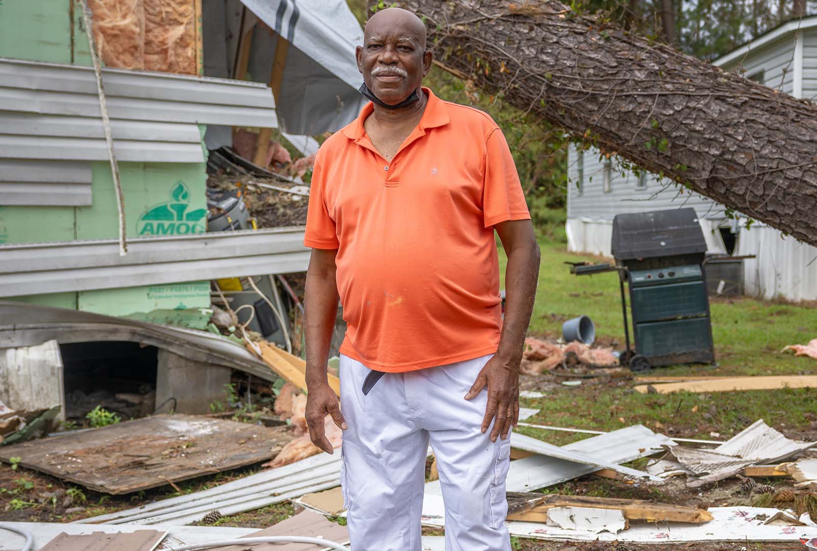 Man stands in front of his mobile home. The home is damaged and has been hit by a fallen tree.