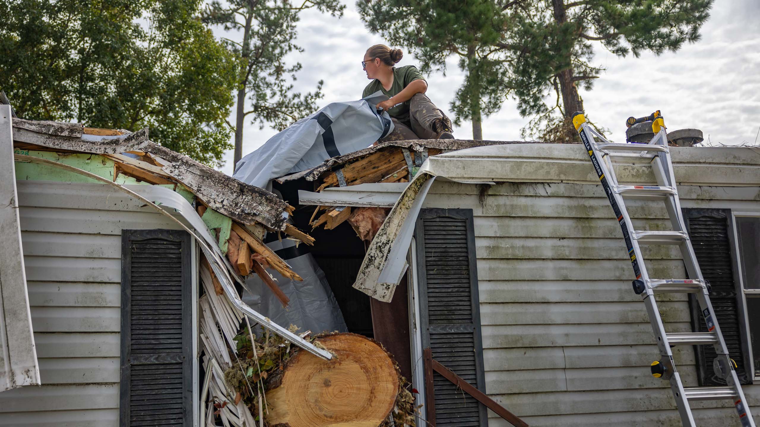 Woman sits on roof positioning a tarp on a destroyed roof where a trunk has slammed through the roof.