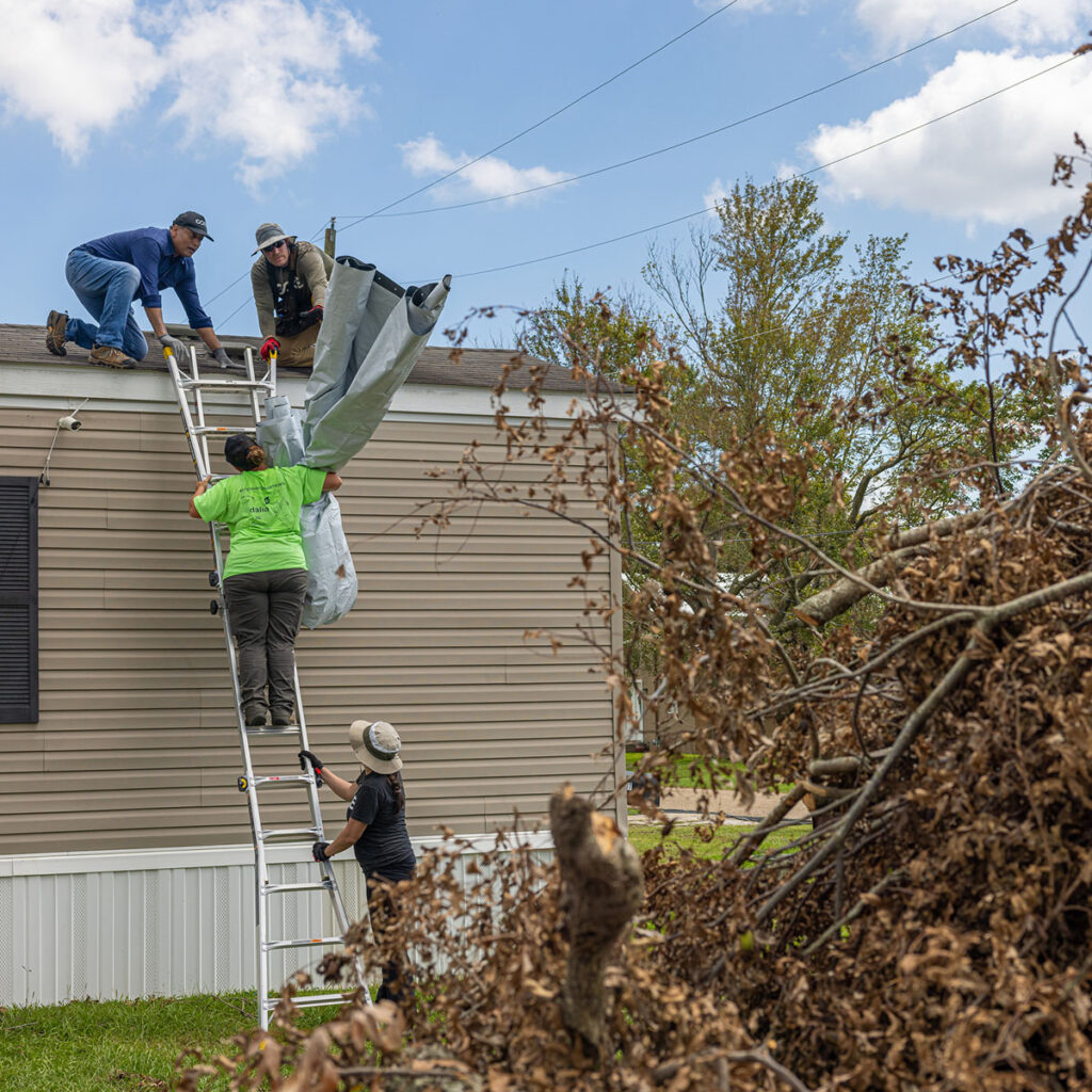 two men on roof of modular home receive a tarp from a woman on a ladder