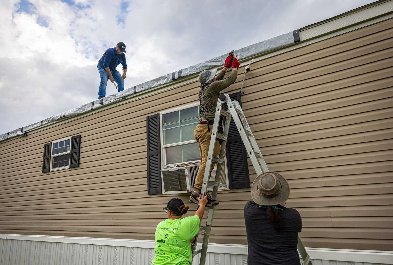 Two people hold ladder against a mobile home. One person stands on ladder to staple tarp. Man on roof bends down to secure tarp