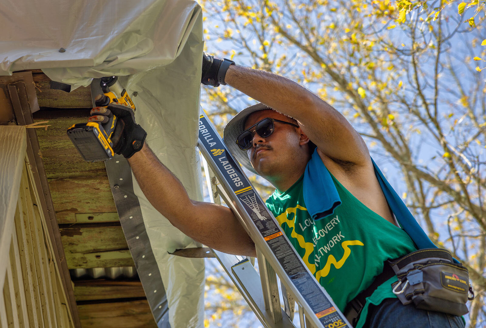 man on ladder leans toward roof to staple tarp